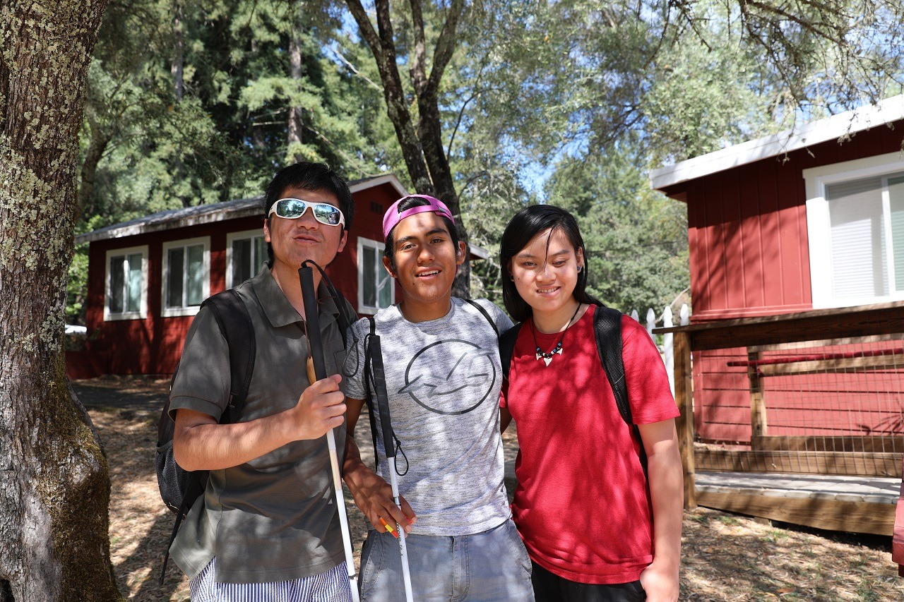 Three teens stand together in front of the lakeside cabins at camp