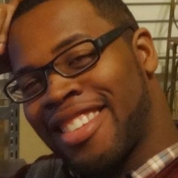 A headshot of Mario, a black man, facing directly into the camera. He is wearing black, rectangular shaped glasses. Mario's right arm is propped on a desk that's out of the shot but his head tilts right and rests on his closed palm. He is wearing a burgundy plaid shirt