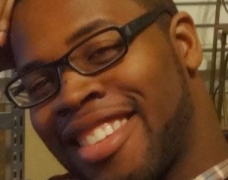 A headshot of Mario, a black man, facing directly into the camera. He is wearing black, rectangular shaped glasses. Mario's right arm is propped on a desk that's out of the shot but his head tilts right and rests on his closed palm. He is wearing a burgundy plaid shirt
