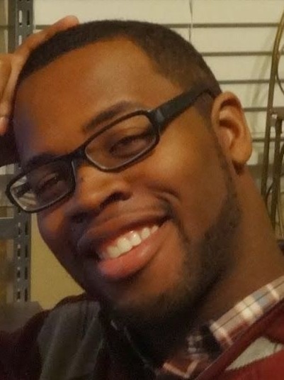 A headshot of Mario, a black man, facing directly into the camera. He is wearing black, rectangular shaped glasses. Mario's right arm is propped on a desk that's out of the shot but his head tilts right and rests on his closed palm. He is wearing a burgundy plaid shirt