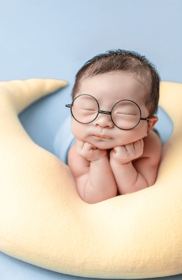 An infant, wearing glasses, who is falling asleep with his head raised, leaning on his arms on top of a moon-shaped pillow
