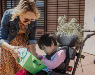 LightHouse Little Learners Specialist, Elizabeth, holds a sensory activity tray filled with beans and colorful plastic bugs for a little girl who is a wheelchair user