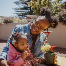 A mother cuddles with her daughter who is playing with a rainbow slinky toy. The mother is smiling and holding onto a silk flower planted in a pot
