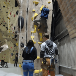 Two LightHouse students wearing rock climbing harnesses watch another student climb the rock wall in front of them