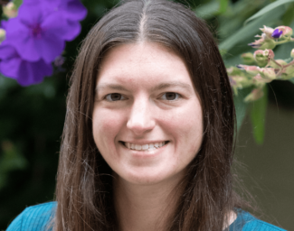 A head shot of Lauren, who has brown shoulder length hair, is wearing a teal colored top and is standing in front of green plants with purple flowers