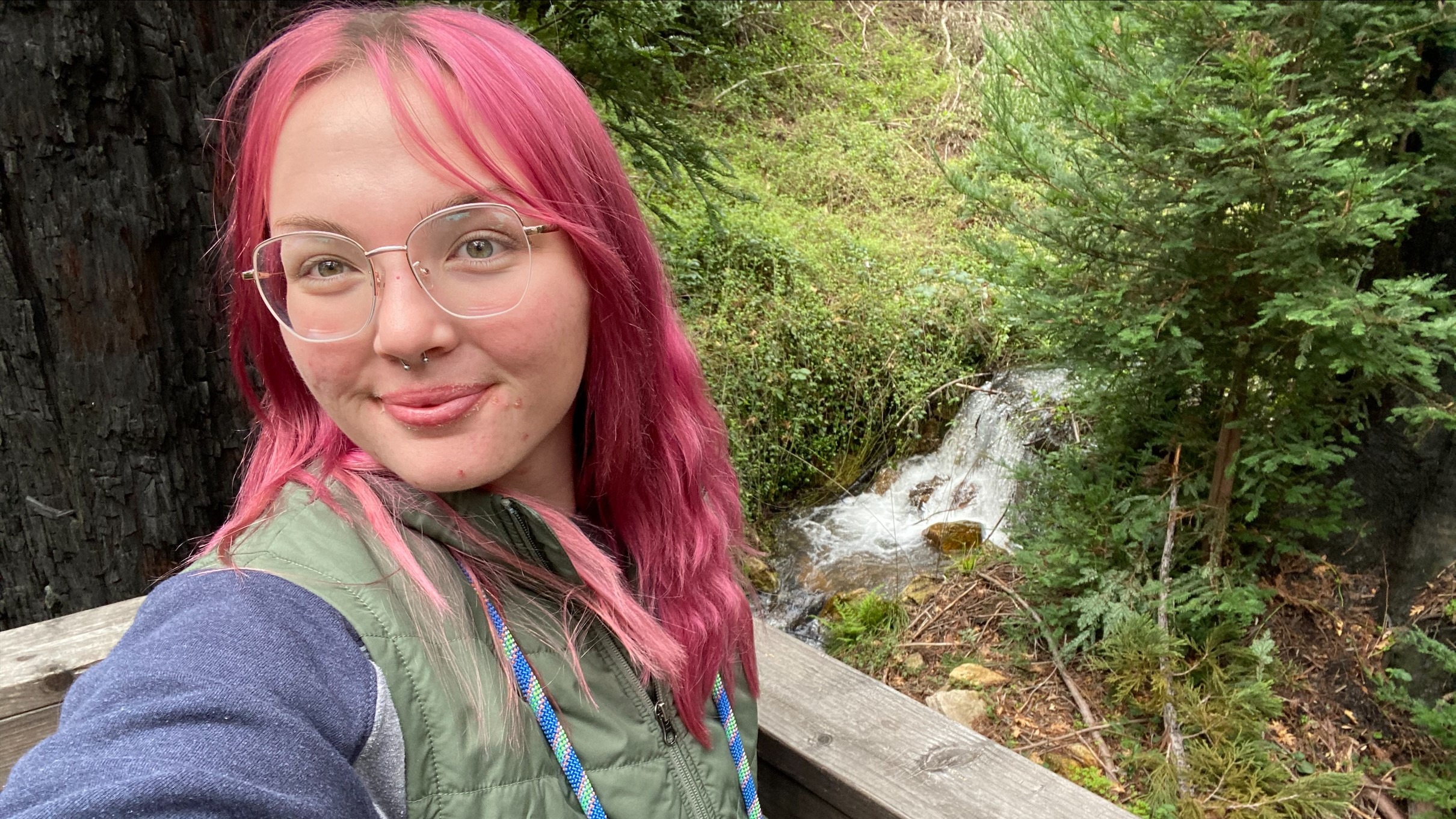 At EHC, Ellie, a young woman with pink hair, takes a selfie outdoors from the corner of a patio with a stream in the background
