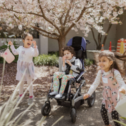 Beeper Egg Hunt ’24 1. Three children smile holding Easter baskets, the child in the middle is a wheelchair user