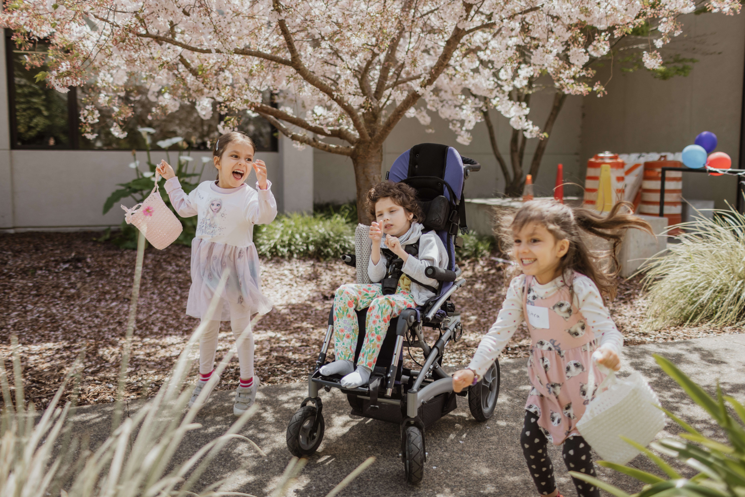 1. Three children smile holding Easter baskets, the child in the middle is a wheelchair user