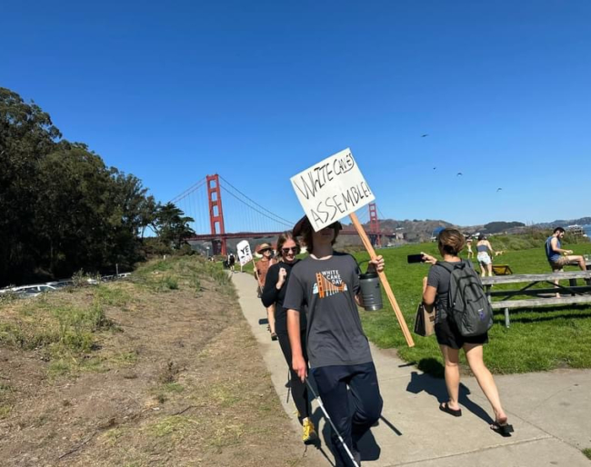 White Cane Day 2024 4 Marchers walk down the dirt path from the Golden gate Bridge to the Chrissy Field after-party. One sign reads, “White Canes Assemble!”