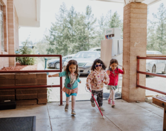 Three Little Learner campers, one using a white cane, run together past the dining hall at Enchanted Hills Camp.