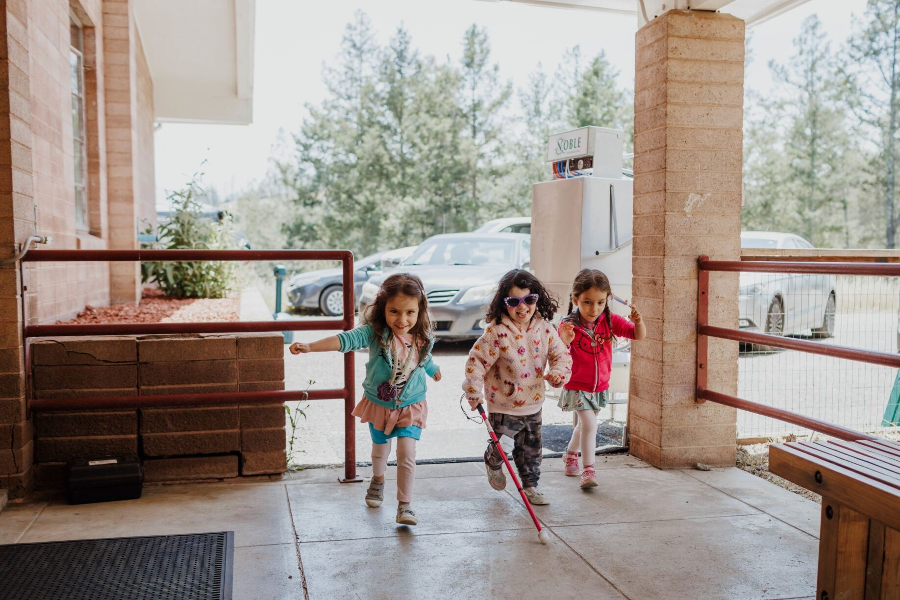 Three Little Learner campers, one using a white cane, run together past the dining hall at Enchanted Hills Camp.