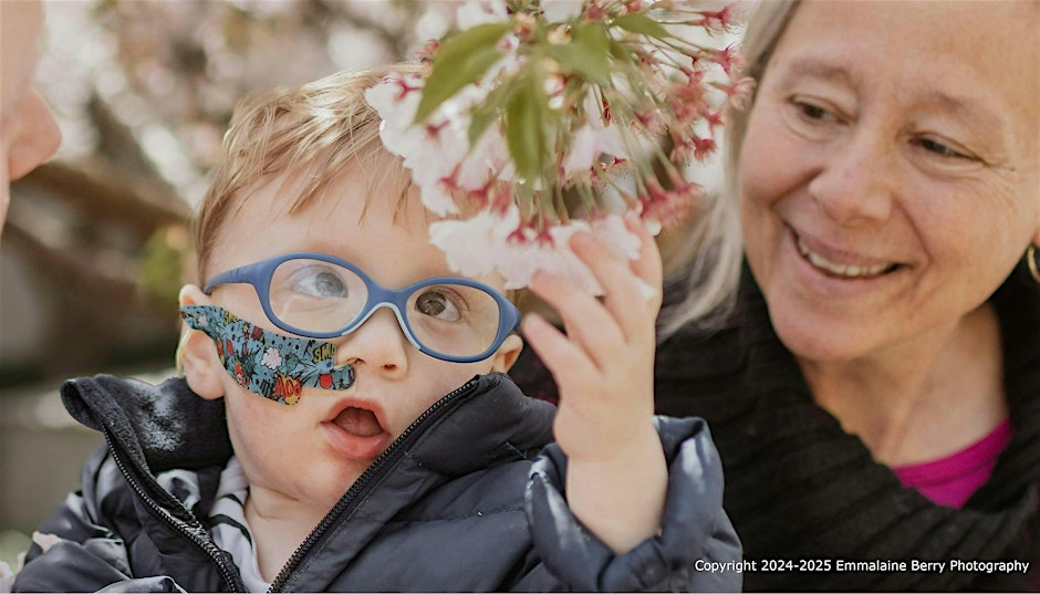 A young child, wearing glasses, reaches out to touch some flowers hanging from a plant as an adult looks on.