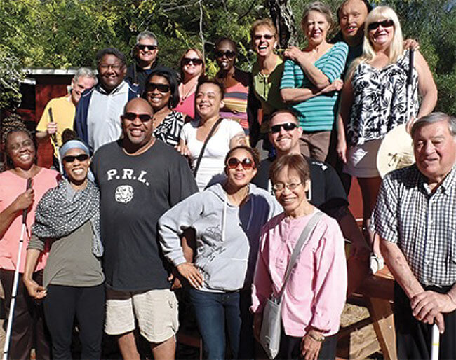 A diverse group of smiling adults stands outside the Enchanted Hills Camp and Retreat cabins, some with white canes, others with dark glasses.