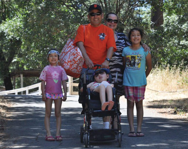 Mom, Dad, outside smiling and walking on a sunny day with their daughters: one child is in a stroller; two sisters are walking next to Mom and Dad.