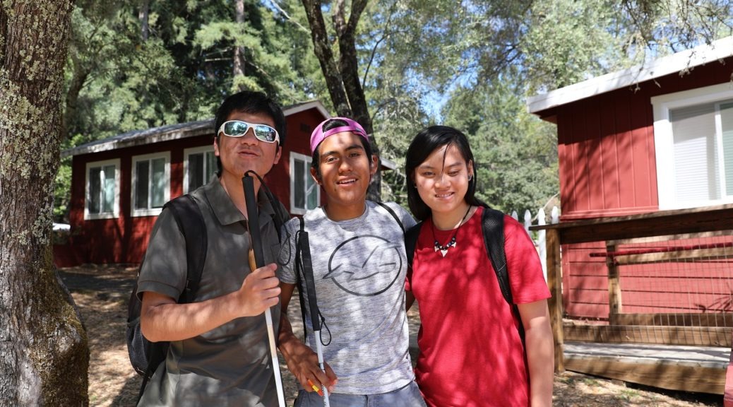 Three teens stand together in front of the lakeside cabins at camp
