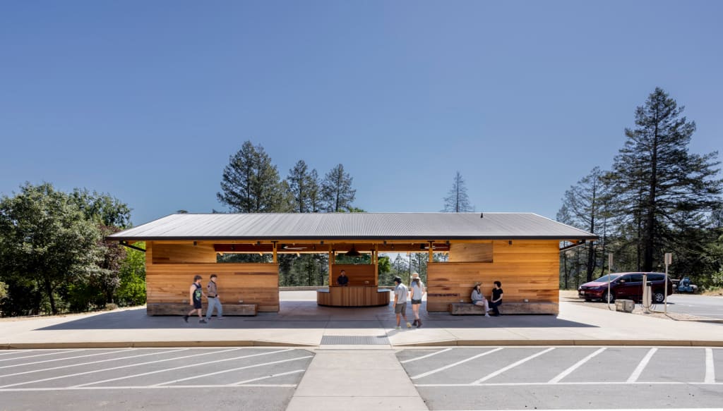 Campers, some with white canes, approach the Welcome Pavilion, a long, gabled redwood-clad open-air pavilion with dark metal roof. Large timber benches, salvaged from the fire, rest under deep awnings and provide shade for campers sitting and chatting on the benches. A large reception desk sits centrally under the pavilion, on axis with the cane-detectable path leading from the parking lot.
