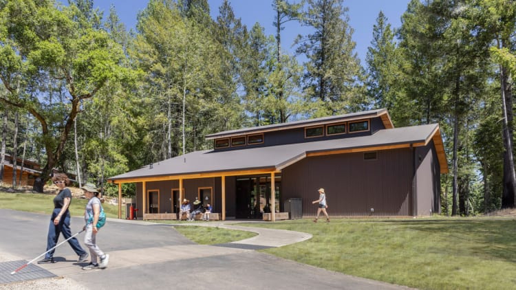 Two campers with white canes cross a cane-detectible path with the Blindness Training Center in the background. The gabled structure, situated with the forest beyond, welcomes campers through an expansive awning with campers chatting on large timber benches underneath. The dark metal-clad structure with large timber columns and redwood eaves evokes the materiality of the charred redwood bark of the trees that survived the fire.