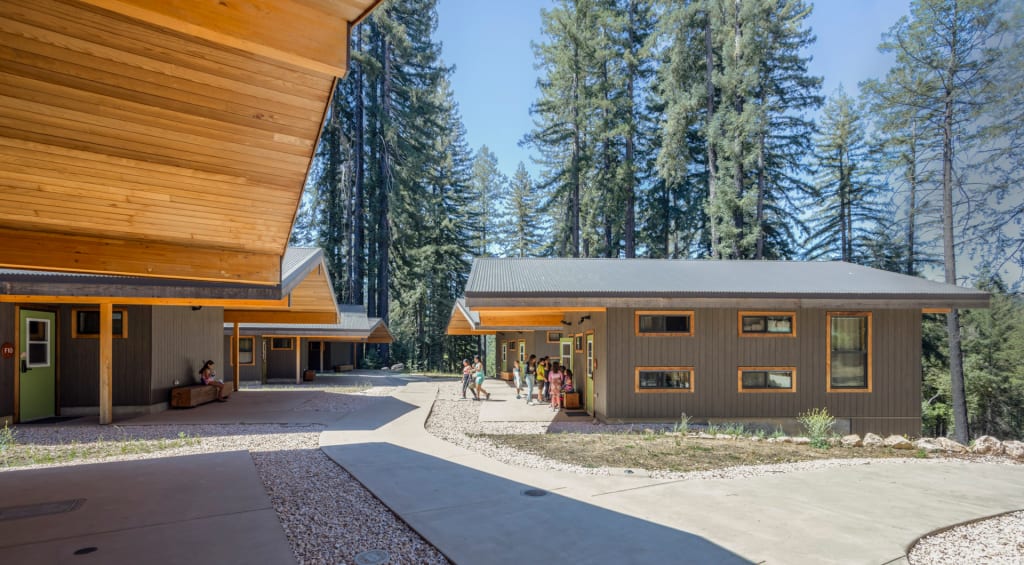 Image of the Forest Family Cabins, taken from underneath the deep redwood eave overhangs of one of the cabins. Youth campers, some with white canes, gather under the awning of a cabin on the right, while a camper with a white can sits in the shade of the opposite cabin. The mature redwood forest provides a lush backdrop to Forest Village.