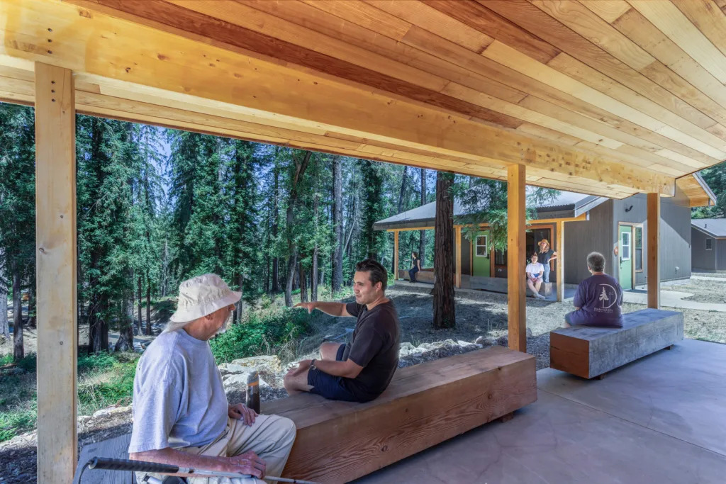 A man with a white cane talks with another man, both seated on a salvaged heavy timber bench, situated under a deep redwood eave overhang on the patio of a Forest Cabin. The patio overlooks redwoods on a hillside to the left, and a paired Forest Cabin with a corresponding porch and eave on the right.