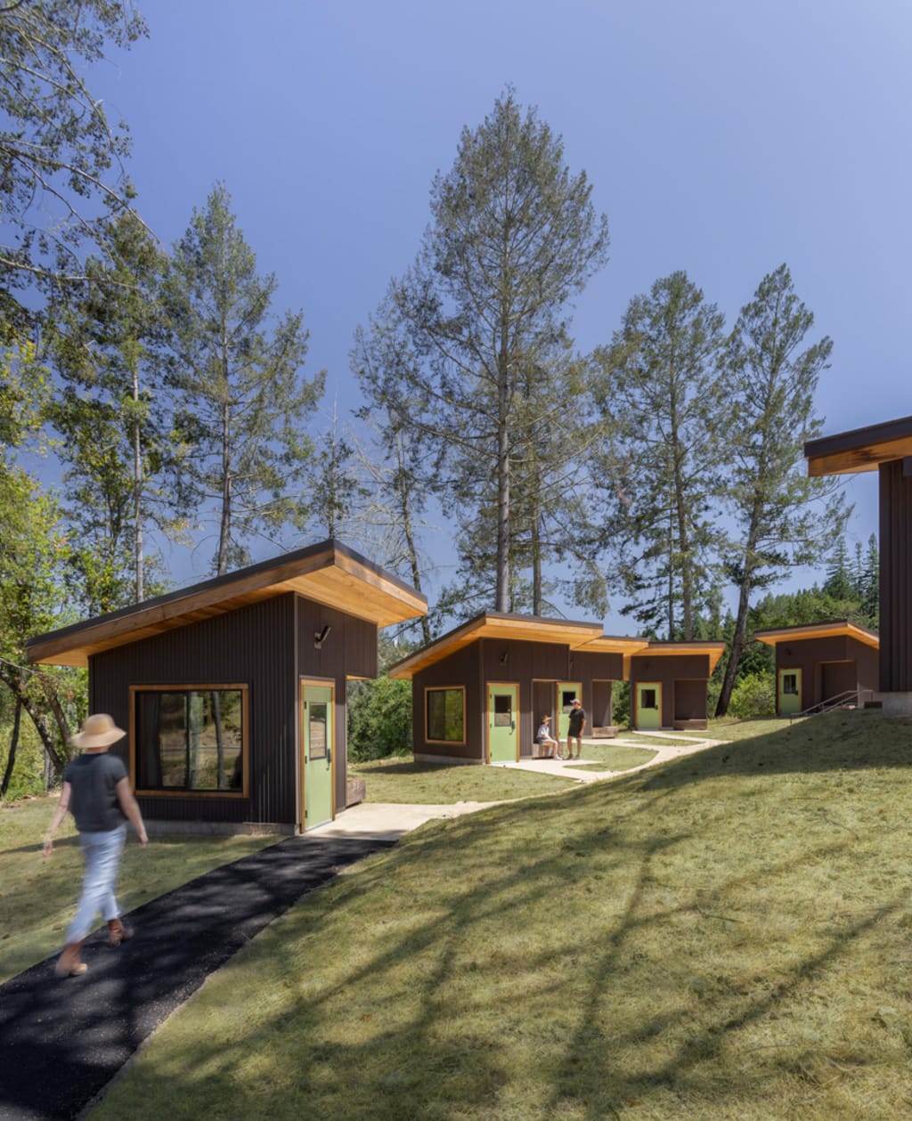 Five Hillside Cabin bungalows with shed roofs, green doors, dark metal cladding, and redwood eaves are situated along a curved cane-detectible path. Trees line the hillside in the distance.