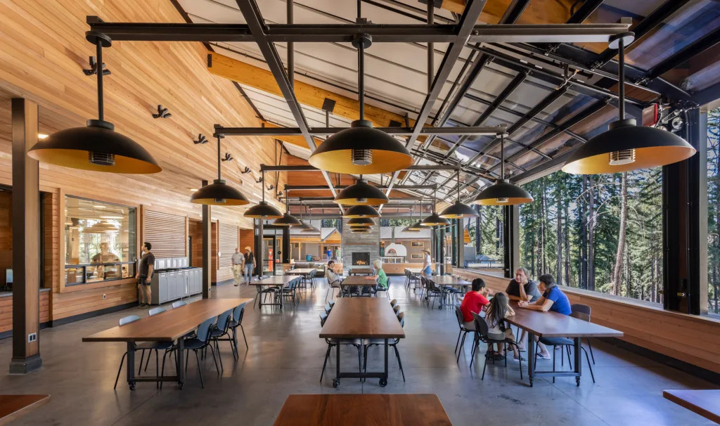 Image of the interior of Forest Commons - an indoor/outdoor dining space with large, glass overhead doors that open up to the redwood forest on the right. A concrete fireplace anchors the space, with radiant heaters and dome light pendants providing light and warmth in the vaulted space. Redwood siding lines the interior walls, with fabric-wrapped acoustic panels on the ceiling, between the heavy timber beams.