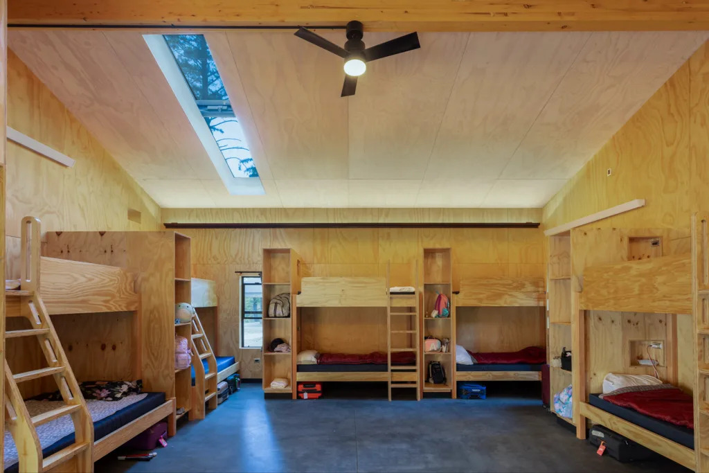 Interior image of a new Forest Bunkhouse Cabin. Cabin interior is clad in plywood, with a whitewashed ceiling, large operable skylight, and ceiling fan. Custom plywood bunkbeds line the perimeter of the space, and offer ample storage for campers' belongings in built-in shelving and niches.
