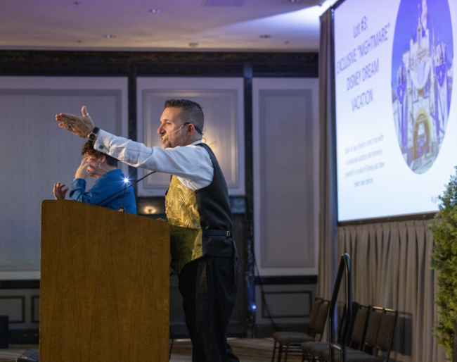 Gala 23 Greg Quiroga Auctioneer Greg Quiroga wears and gold vest and stands at the podium talking excitedly with his hands in the air