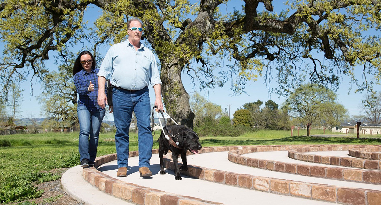 Woman helping man walking through labyrinth with assistant dog.